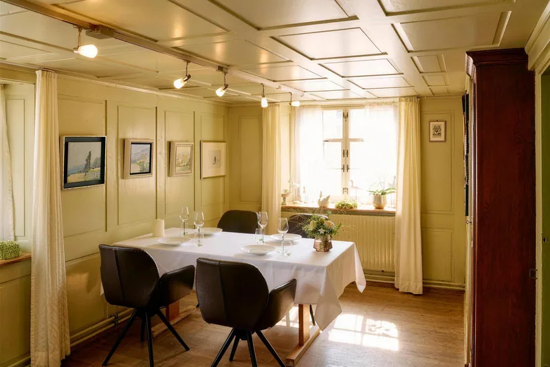 Eine helle Stube mit Täfer, Bildern an der Wand und einem gedeckten Tisch am Fenster